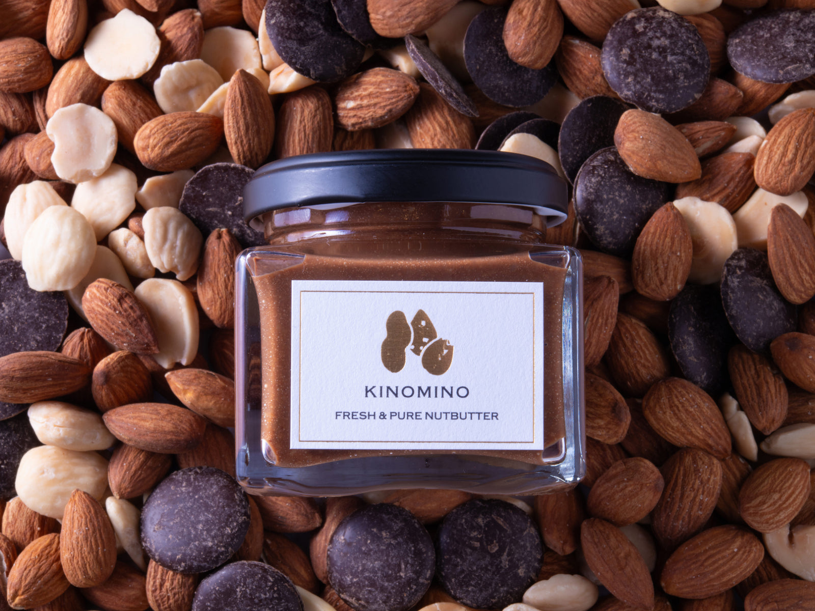沢山のアーモンドとクーベルチュールチョコレートを背景にした、チョコレートアーモンドバターの商品写真